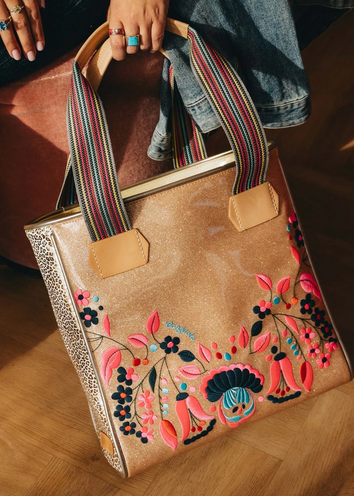 Brown tote bag with colorful floral embroidery held by a person on a wooden floor.